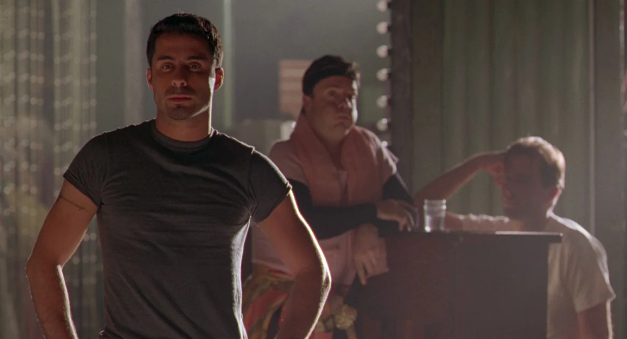 The Birdcage (1996) movie still: medium — A dimly lit room with mirrors and light fixtures along the edges. A muscular man stands in the foreground…