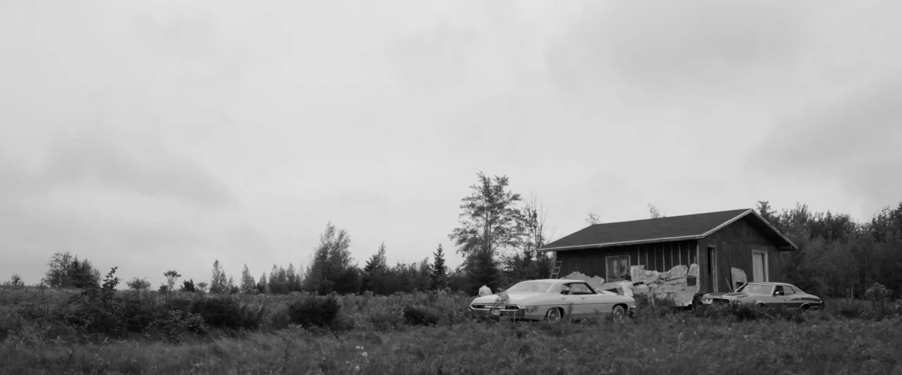 Weirdos (2016) movie still: wide — An overgrown field contains a small, weathered wooden house with peeling siding and two vintage cars. A large…