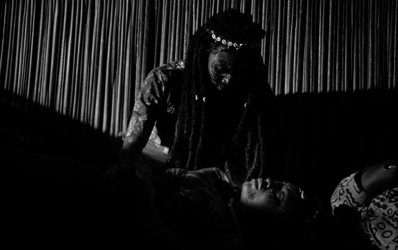 Mami Wata (2023) movie still: medium — A dark interior space with walls made of vertical wooden reeds or bamboo. One woman leans over another woman…