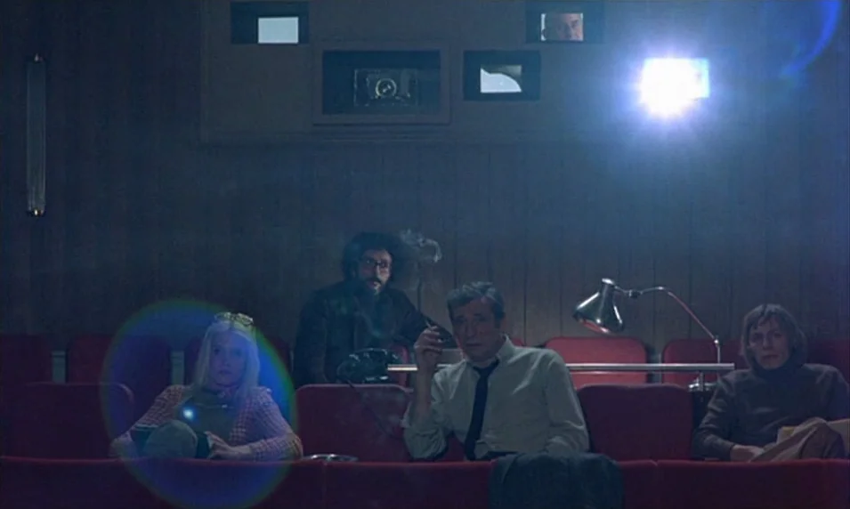 Tout Va Bien (1972) movie still: wide — A dark screening room features red padded chairs arranged in a row. Four people sit in the chairs while a…