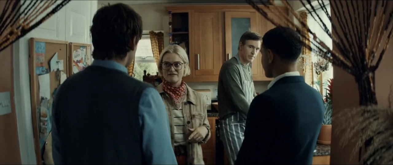 Bonus Track (2024) movie still: medium — A residential kitchen features light wood cabinets and a cork board on the wall. Three people stand in the…