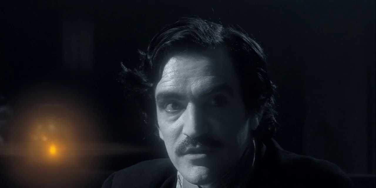Twixt (2011) movie still: closeup — A dark, minimally lit room frames a man's face in deep shadow. He stares off-screen while a single bright orange…