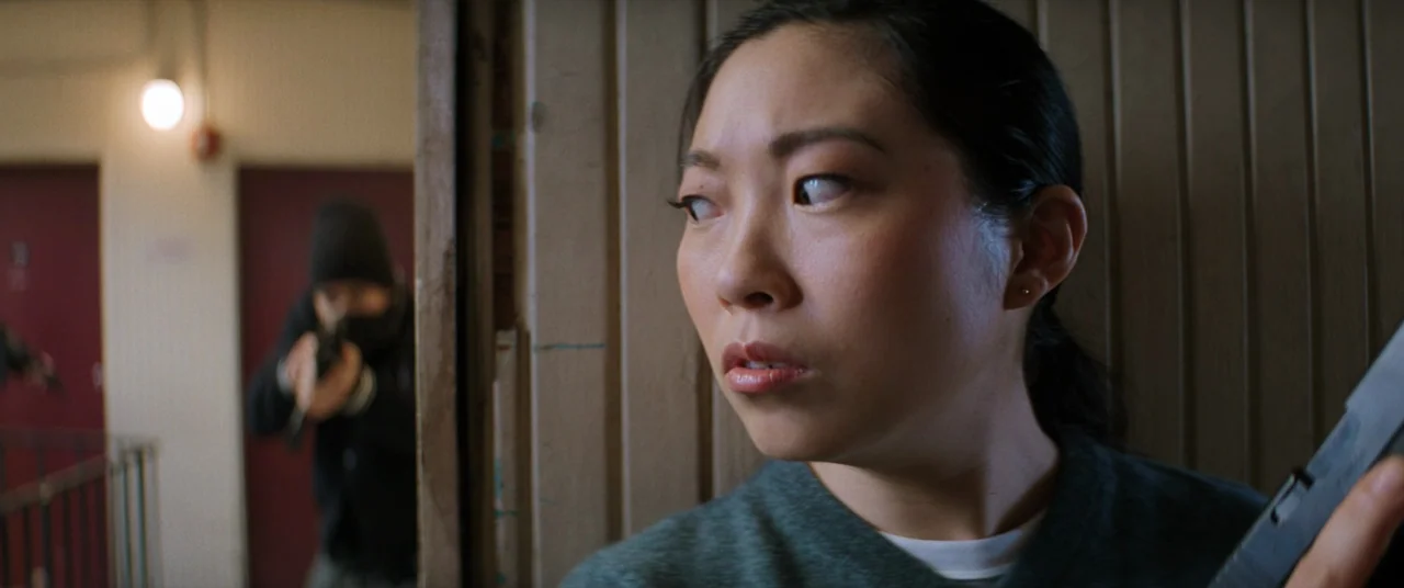 Renfield (2023) movie still: closeup — A dimly lit residential hallway features vertical wood paneling and closed red doors. A young woman peers…