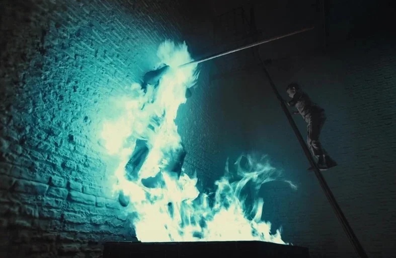 Gretel & Hansel (2020) movie still: wide — An industrial room with exposed brick walls is illuminated by a source of blue fire. A man hangs from a…