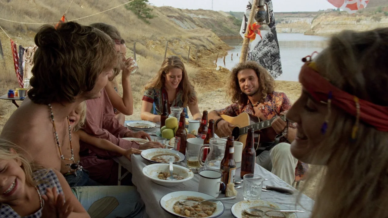 The Sect (1991) movie still: medium — A group of young adults shares a meal at a long table by a quarry. The man plays a guitar while the others eat…