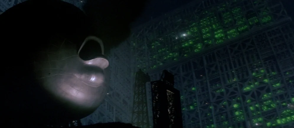 Dune (1984) movie still: establishing — A massive industrial structure looms in the darkness with glowing green lattice frameworks. A large metallic…