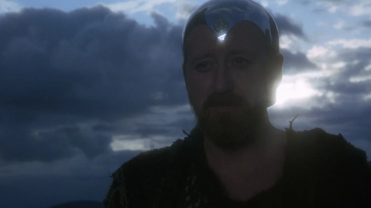 Excalibur (1981) movie still: closeup — An outdoor landscape under a cloudy dusk sky serves as the background. A man wears a metallic headpiece and…