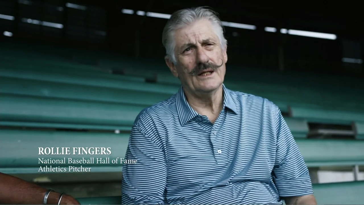 Reggie (2023) movie still: medium — An outdoor seating area with rows of green wooden bench rows. A man with a distinctive mustache sits in the…