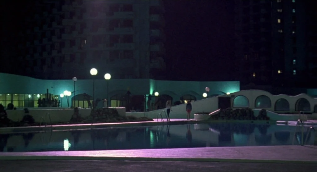 Morvern Callar (2002) movie still: wide — A night view shows a commercial pool area with geometric architecture and high-rise hotel structures. Two…