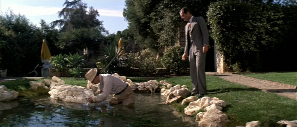 Chinatown (1974) movie still: wide — A man in a suit stands by a stone-lined pond in a manicured lawn. A second man wears a hat and kneels in the…