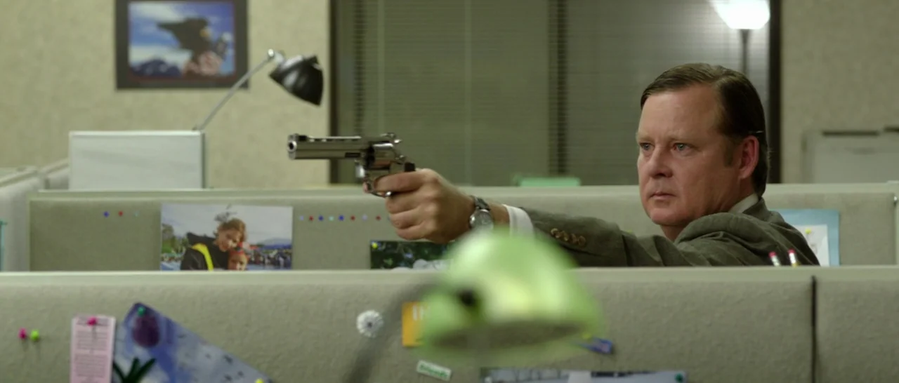 God Bless America (2011) movie still: medium — A sterile workplace cubicle features beige fabric partitions decorated with personal photographs and…