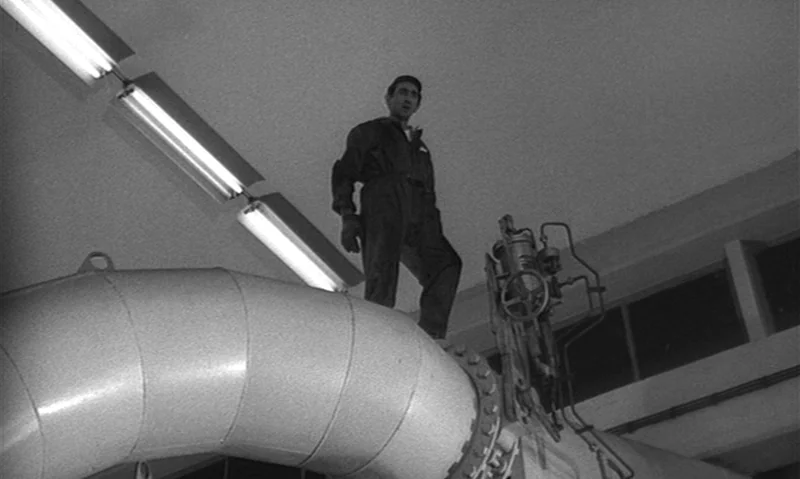 Man Is Not Bird (1965) movie still: fullbody — An industrial facility interior featuring exposed concrete walls and large metal ventilation pipes. A…