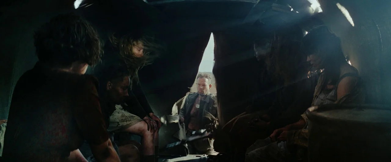Sisu (2022) movie still: wide — A dark, cramped interior of a transport vehicle with dusty surfaces and dim lighting. Several people sit in shadow…