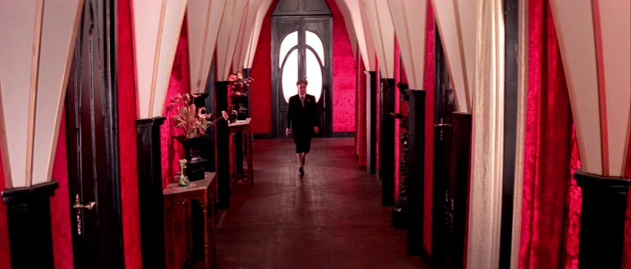 Suspiria (1977) movie still: wide — A long symmetrical corridor features bright red walls and white vaulted arching ceiling supports. A man in a…