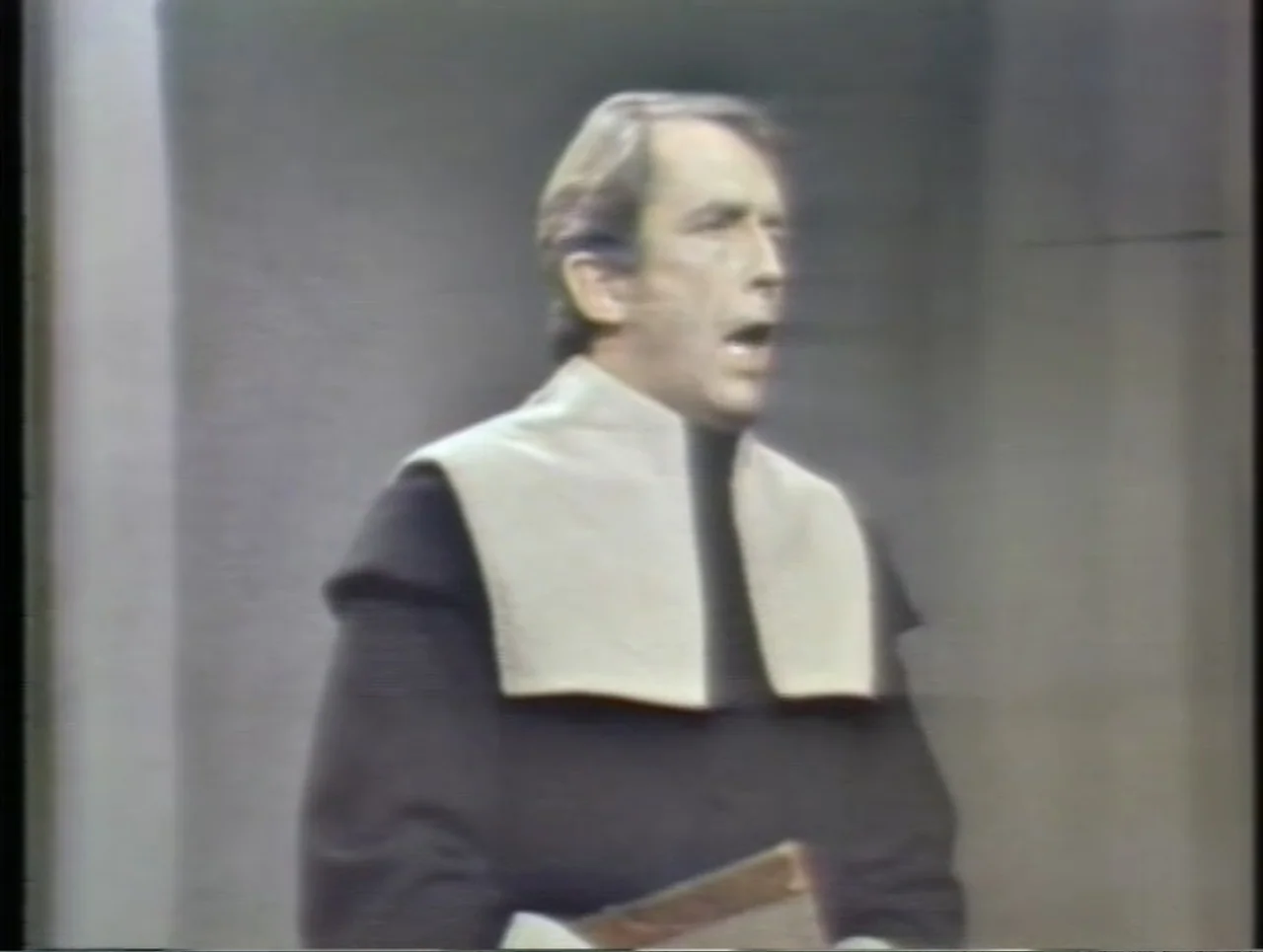 The Crucible (1996) movie still: medium — A man stands in front of a plain wall resembling a stone interior. The man wears a dark robe and speaks…