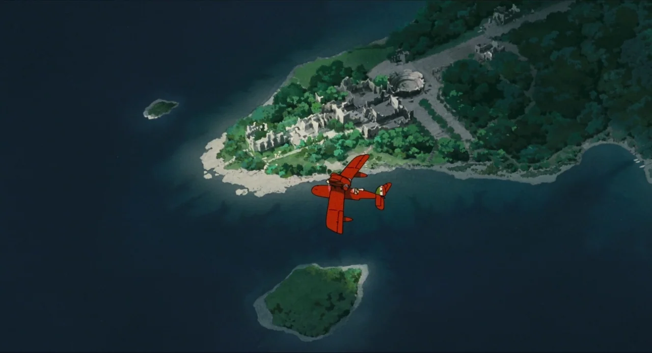 Porco Rosso (1992) movie still: wide — A top-down view shows a red seaplane flying over dark water near a coastline. The coast features dense green…
