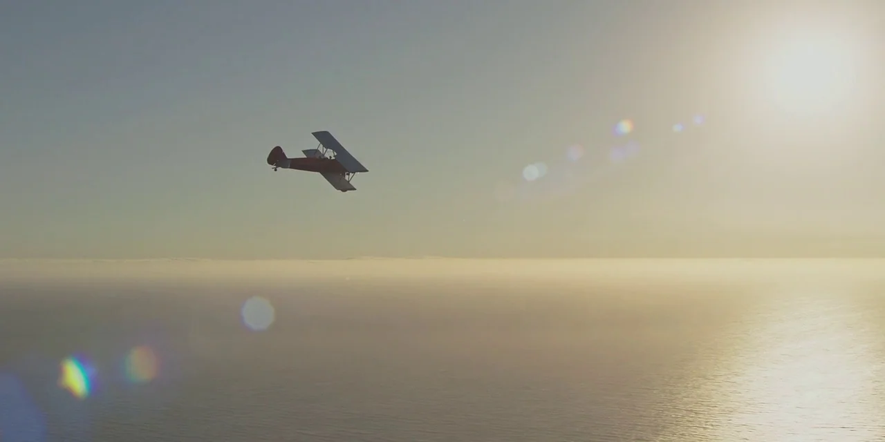20th Century Women (2016) movie still: wide — A vintage red biplane cruises over a vast expanse of ocean at sunset. The bright sun hangs in the sky…