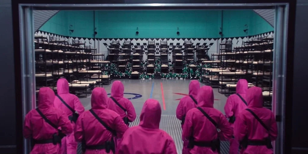 Squid Game (2024) movie still: wide — A large industrial hall features tiered metal bunk beds lining the teal walls. Guards in bright magenta…