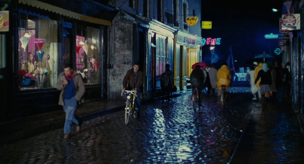 The Umbrellas Of Cherbourg (1964) movie still: wide — A rain-slicked cobblestone street at night is lined with shop windows glowing with light.…