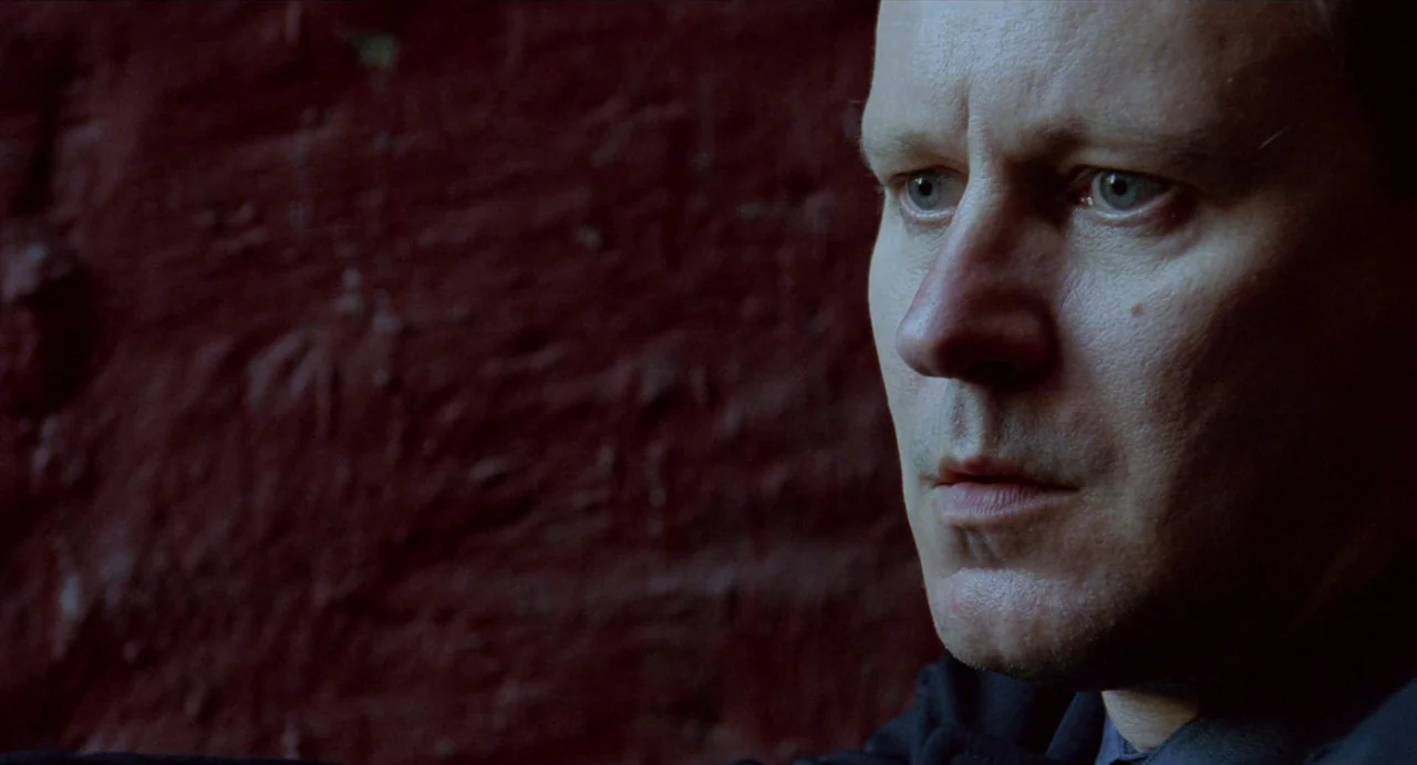 Insomnia (1997) movie still: closeup — A man stands before a textured, deep red wall surface. He stares forward while remaining still against the…