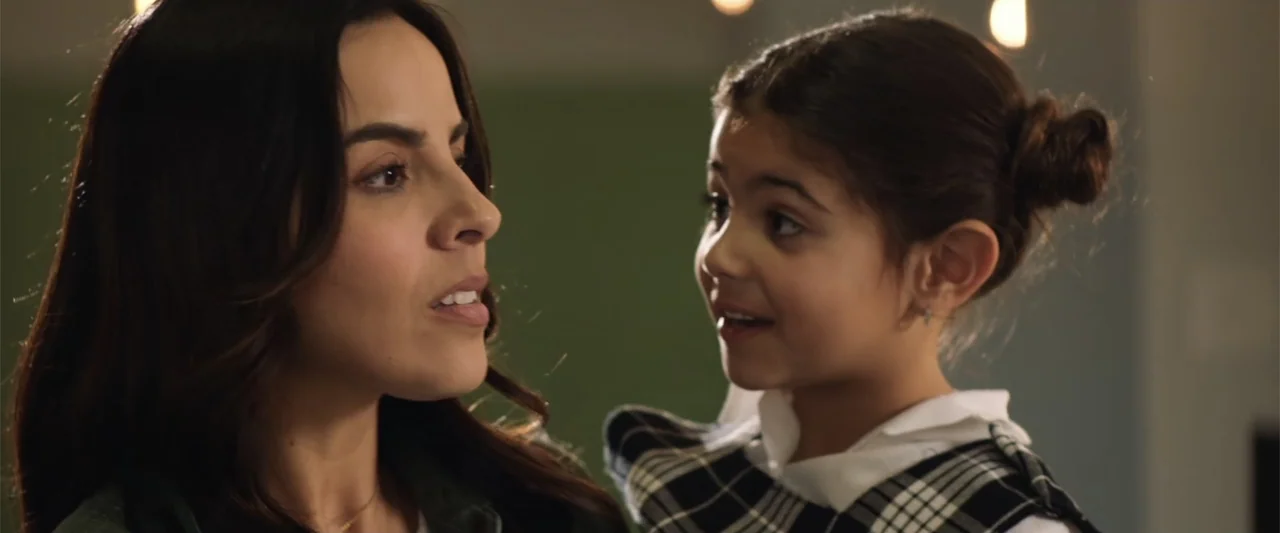 Winner Takes the Cake (2025) movie still: medium — A woman and a young girl stand close together inside a brightly lit room. They face each other…