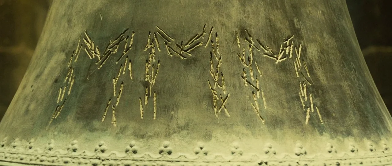 A Very Long Engagement (2004) movie still: closeup — A close-up view of a metallic bell surface with engraved runic markings. The surface shows…