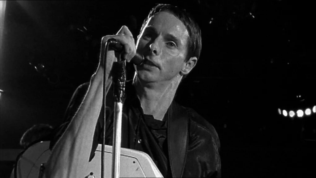 24 Hour Party People movie still: medium — A dimly lit concert venue with black background shadows behind the musician. A man holds a microphone…