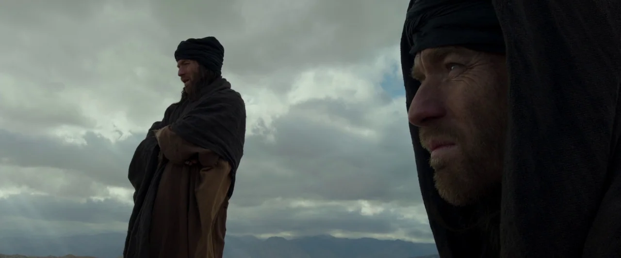 Last Days in the Desert (2015) movie still: medium — A desolate desert landscape beneath a heavy, overcast grey sky. Two men in dark traditional…