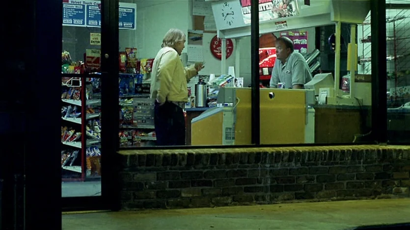 Love Liza (2002) movie still: wide — A brightly lit convenience store interior is seen at night through a glass window. A man stands at the counter…
