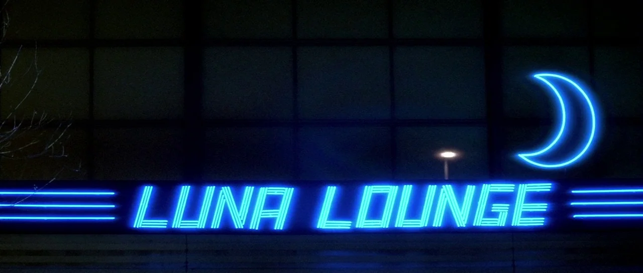 Lost Highway (1997) movie still: wide — A dark, grid-patterned exterior wall features a prominent glowing blue neon sign. The sign displays the text…