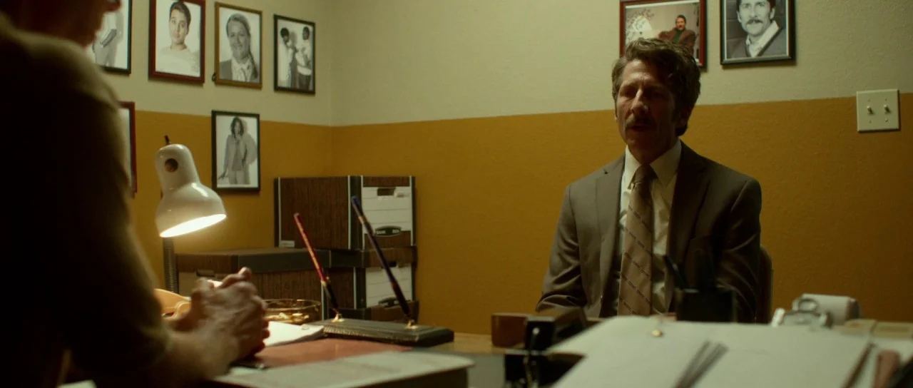 Faults (2014) movie still: medium — A drab room with mustard-colored walls contains several framed photographs hung in rows. A man in a brown suit…