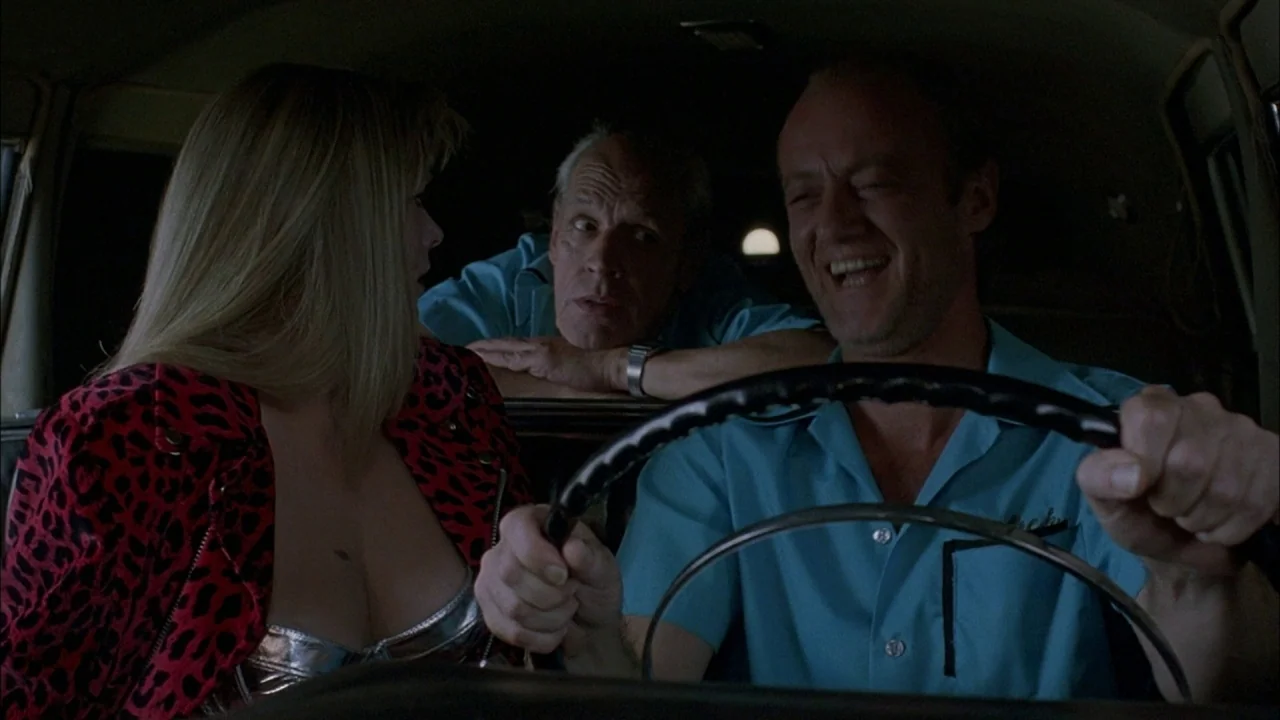 Whore (1991) movie still: medium — The interior of a moving van at night contains three people. The driver smiles while the other two passengers…