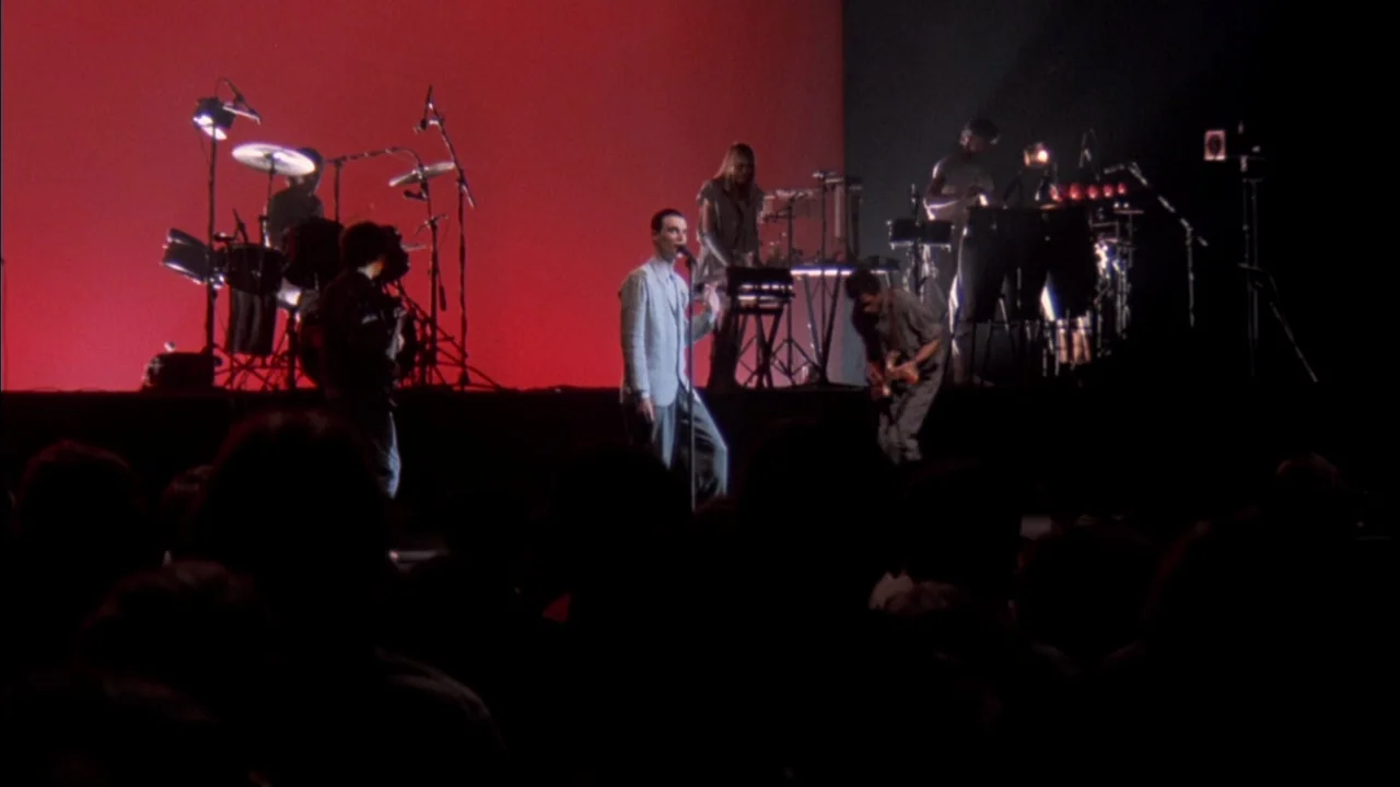 Stop Making Sense (1984) movie still: wide — A musical performance occurs on a sparse, minimalist stage with a solid red backdrop. The band members…