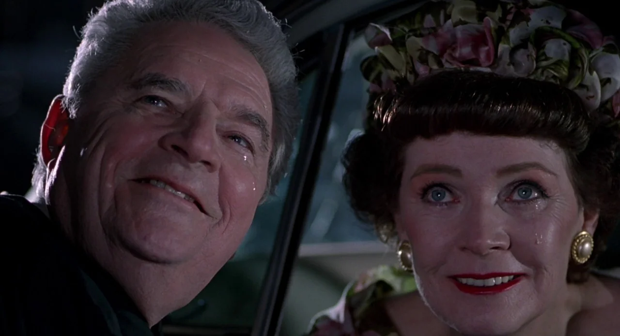 Cry Baby (1990) movie still: closeup — Inside a vehicle at night, two older adults look forward through a window. Both individuals exhibit visible…