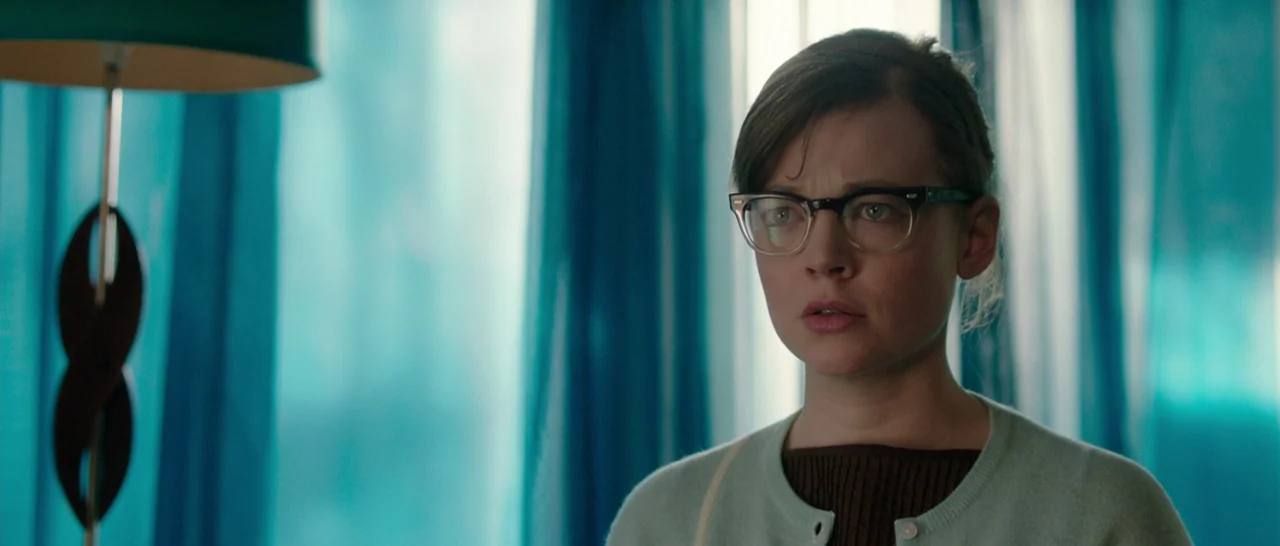 Predestination (2014) movie still: medium — A woman stands indoors in front of large, bright teal sheer curtains. She stares forward with a neutral,…