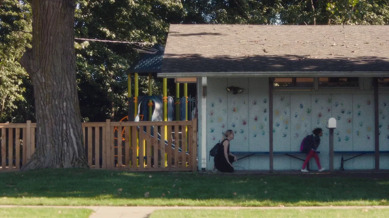 Saint Frances (2019) movie still: wide — A light-colored building wall covered in colorful handprint paintings faces a wooden picket fence. An adult…