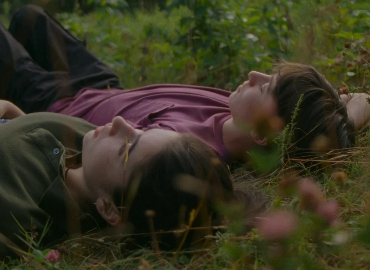 Falcon Lake (2022) movie still: medium — Two individuals lie side by side on a grassy ground covered with small wildflowers. They lie on their sides…