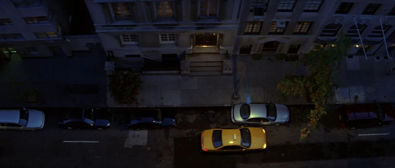 The Thomas Crown Affair (1999) movie still: establishing — A high-angle view of a city street at night showing rows of parked cars. The sidewalk…