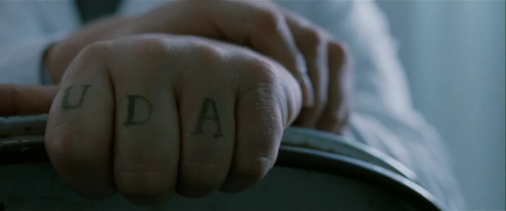 Hunger (2008) movie still: closeup — A close view of a human hand resting on a curved edge. The knuckles are tattooed with the black letters U, D,…