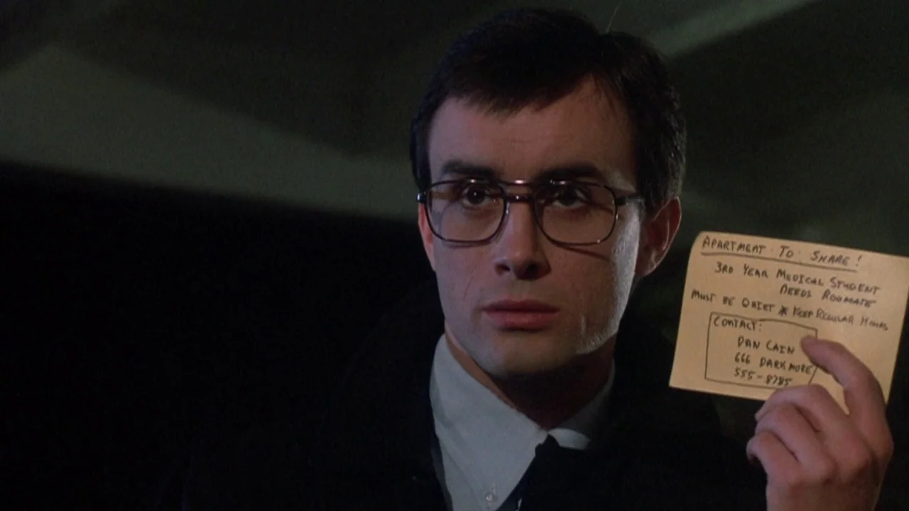 Re-Animator (1985) movie still: closeup — A dimly lit interior space with a dark background and low visibility. A young man wearing glasses holds a…