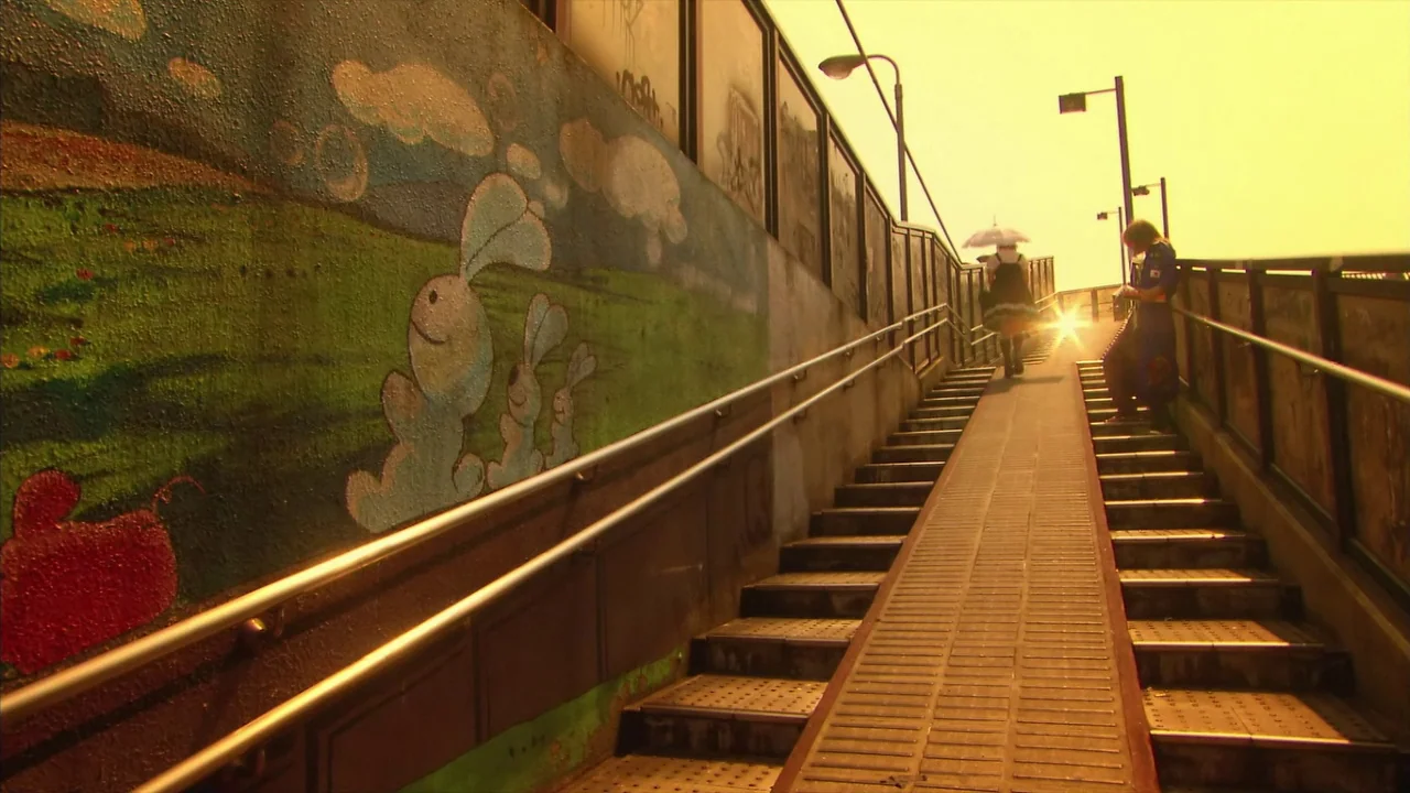 Kamikaze Girls (2004) movie still: wide — An outdoor pedestrian overpass features a ramp flanked by stairs and a wall mural. Two young people walk…