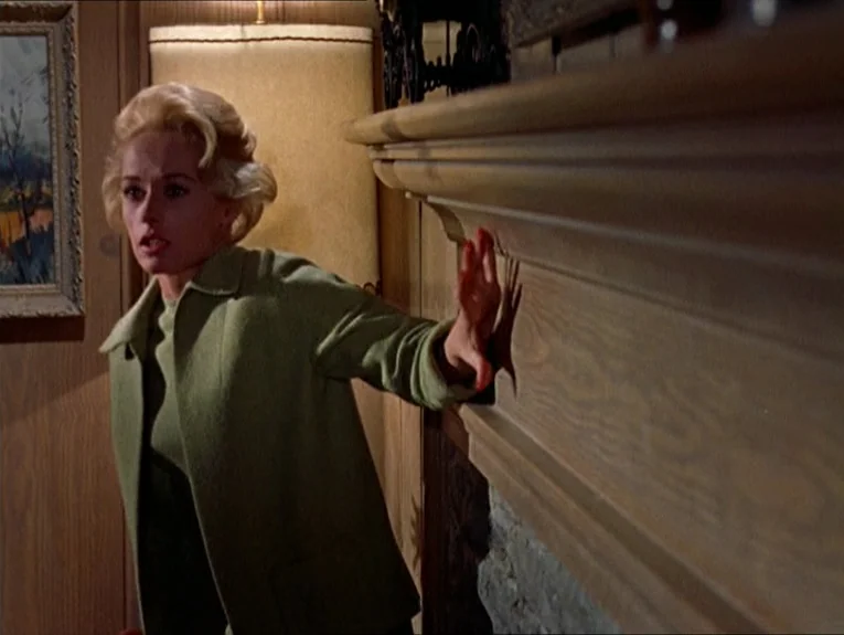 The Birds movie still: medium — A 1960s living room features wood-paneled walls and a stone fireplace mantel. A blonde woman in a green cardigan…