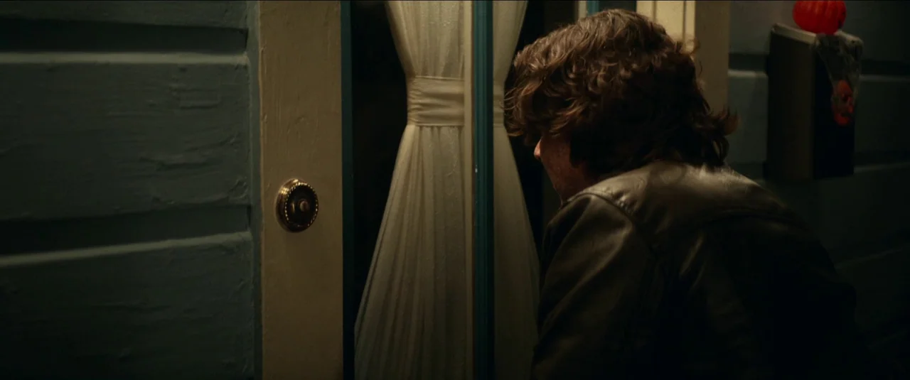 Halloween Kills (2021) movie still: medium — A residential house exterior at night features blue siding and a door with curtains. A man in a dark…