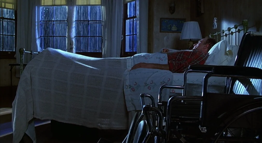 Misery (1990) movie still: wide — A bedroom at night features a metal-framed bed and a parked wheelchair in the foreground. An elderly man rests…