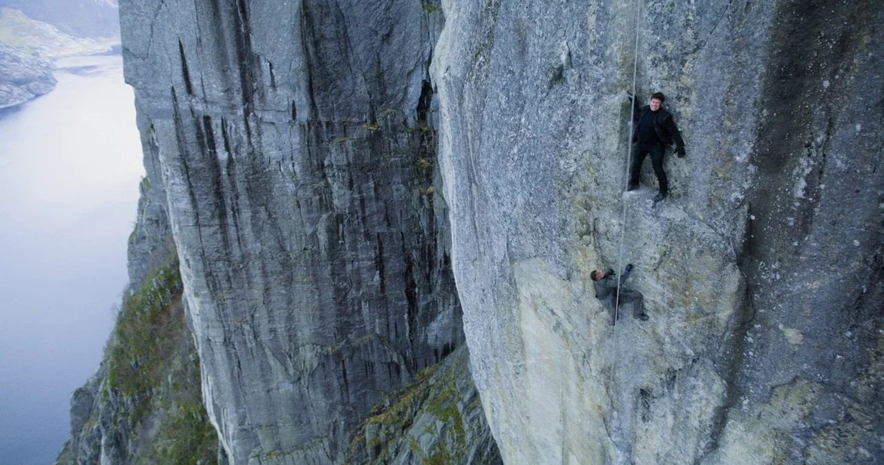 A massive vertical cliff wall rises above a deep blue body of water. Two men are anchored to the rock face by a single vertical rope.