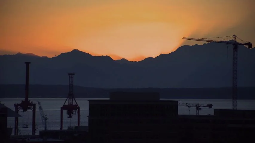 Touchy Feely (2013) movie still: establishing — A harbor cityscape silhouette is set against a vibrant orange sunset over distant mountains.…