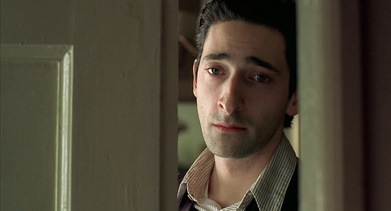 The Pianist (2002) movie still: closeup — A man stands partially obscured by a wooden doorframe in an indoor space. He looks through the opening…
