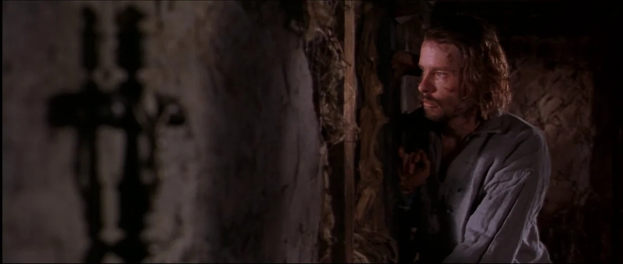 Ravenous (1999) movie still: medium — A dimly lit, cramped interior features textured, mud-plastered walls and raw wooden supports. A bearded man in…