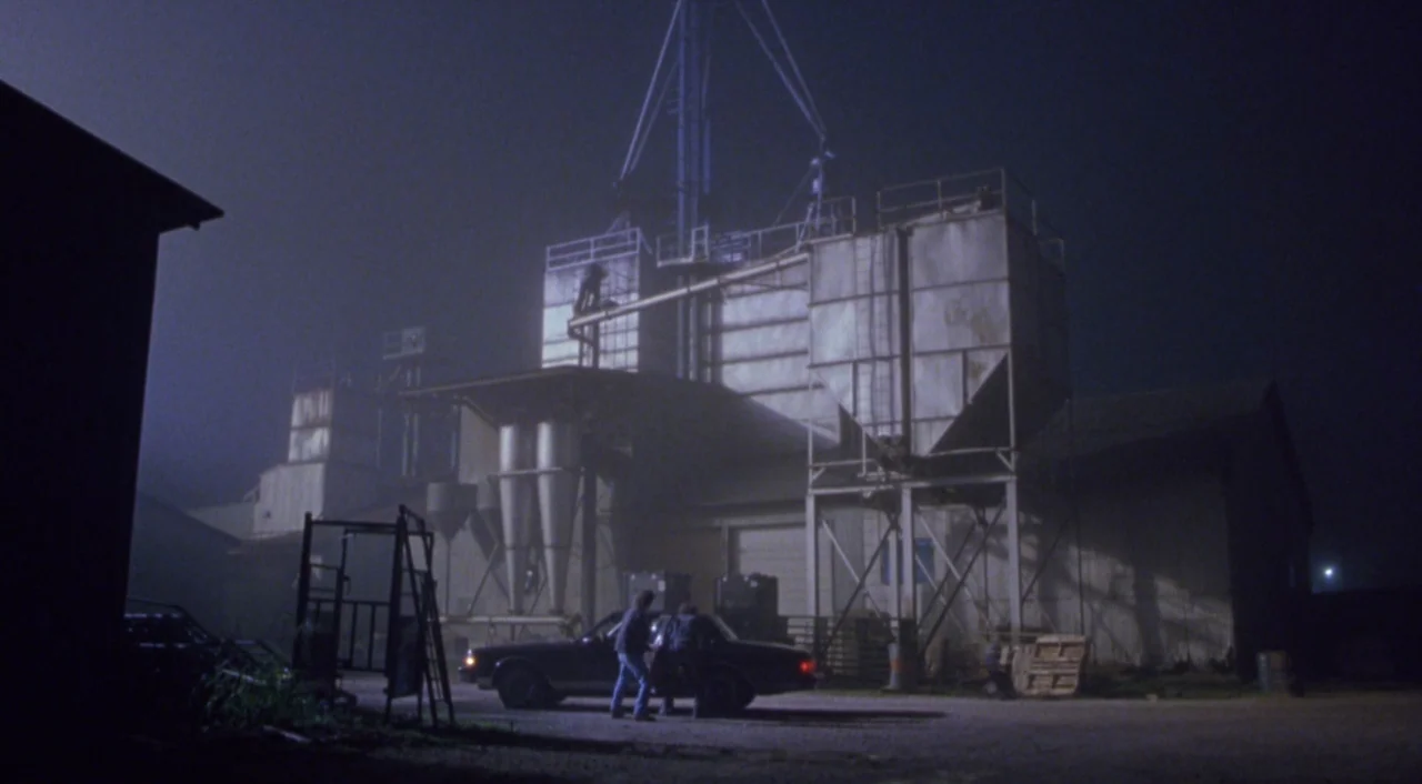 Boys Don’t Cry (1999) movie still: wide — A large industrial facility with tall metal silos stands against a dark, foggy night sky. Two individuals…
