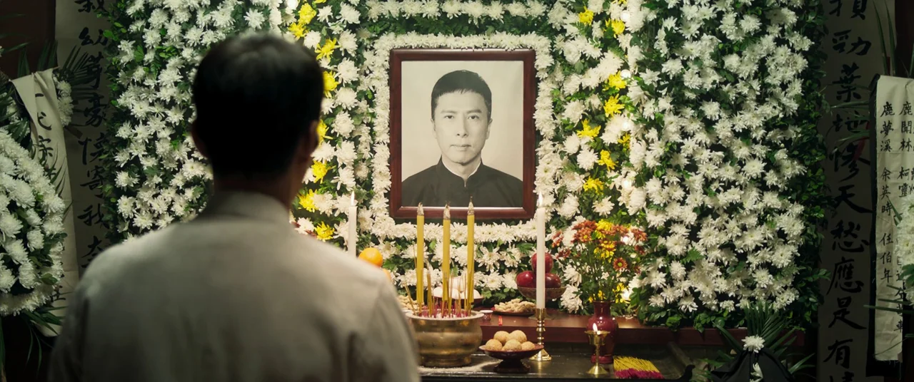 Ip Man 4: The Finale (2019) movie still: medium — A wall is covered in dense white and yellow chrysanthemum flowers framing a portrait. A man stands…
